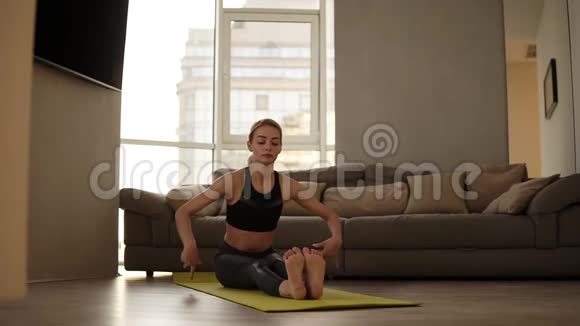 苗条的金发女人坐在垫子上早上在明亮的客厅里靠身体向前伸展运动视频的预览图