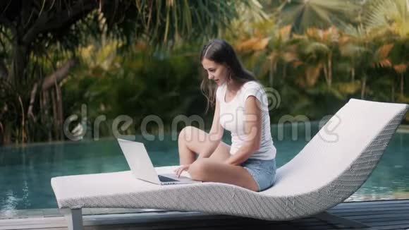 欢乐女孩的侧景在笔记本电脑上工作坐在游泳池附近的沙发上视频的预览图