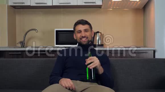 一个穿着深蓝色套衫和灰色裤子的年轻人坐在沙发上喝着啤酒微笑着看电视上的娱乐节目视频的预览图