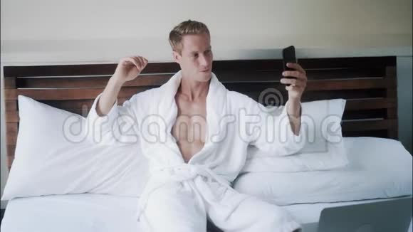 穿着白色浴袍的金发帅哥在床上用手机拍自拍照片视频的预览图