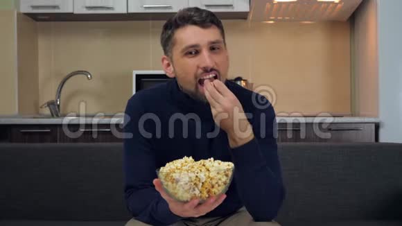 男人坐在沙发上吃透明玻璃盘子里的爆米花微笑着看电视上的娱乐节目视频的预览图