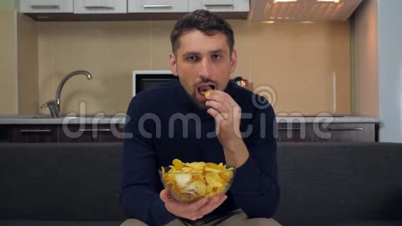一个年轻人坐在沙发上吃透明玻璃盘子里的薯片看电视上的娱乐节目视频的预览图