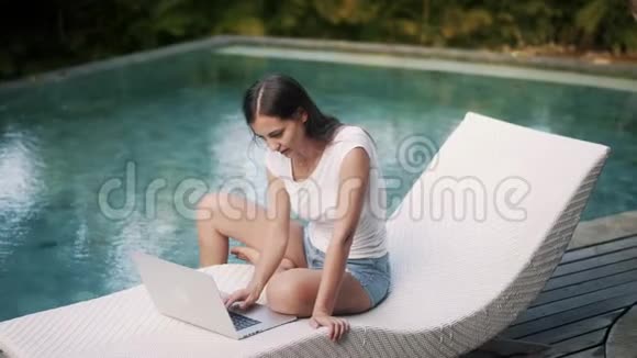 女性自由职业者坐在游泳池附近的躺椅上工作在笔记本电脑上类型在键盘上视频的预览图