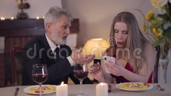 高级优雅的白种人男子西装革履用智能手机与年轻女子交谈穿着红裙子的忙碌女孩无视年长者视频的预览图