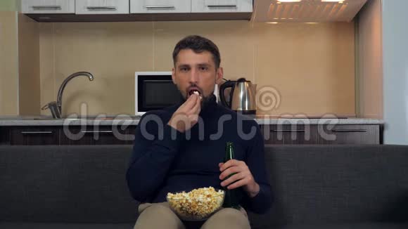 一个穿着深蓝色套衫和灰色裤子的年轻人坐在沙发上吃爆米花喝啤酒看电视上的娱乐节目视频的预览图