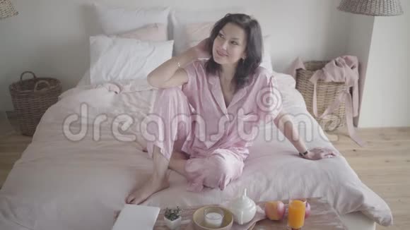 一幅快乐的白种人女人穿着睡衣做梦坐在家里的床上微笑着的黑发女人视频的预览图