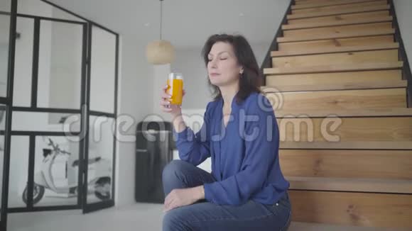 有魅力的白种人女人坐在楼梯上在室内喝橙汁的肖像优雅自信的黑发视频的预览图