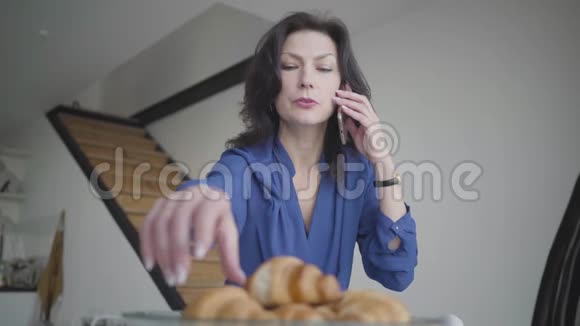 成功的严肃的高加索女人选择牛角面包并在家里打电话的肖像美丽自信视频的预览图