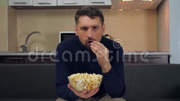 严肃的男人坐在沙发上吃透明玻璃盘子里的爆米花生气在电视节目上发誓视频的预览图