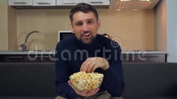 一个年轻人坐在沙发上吃透明玻璃盘子里的爆米花微笑着看电视上的娱乐节目视频的预览图