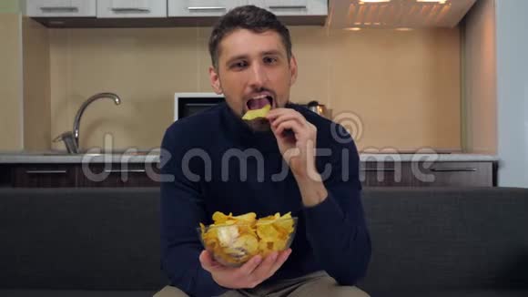 男人坐在沙发上吃透明玻璃盘子里的薯片微笑着看电视上的娱乐节目视频的预览图