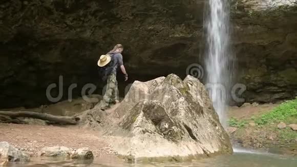 背着背包的年轻人爬上石头看着美丽的瀑布景色视频的预览图