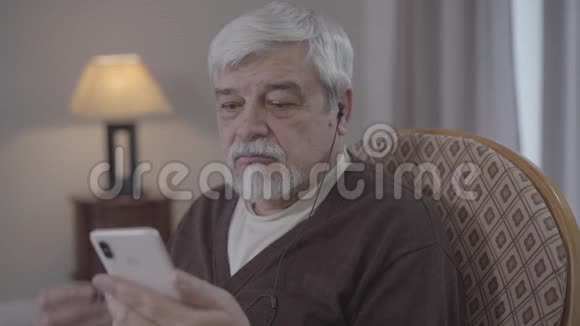 成熟的高加索男人的特写戴上耳机打开智能手机上的音乐正高级退休人员视频的预览图