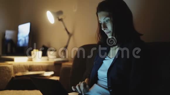 年轻漂亮的女人晚上坐在家里的教练上使用笔记本电脑和浏览社交媒体视频的预览图