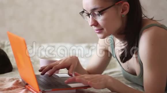 年轻漂亮的女孩正在用她的笔记本电脑工作躺在家里的床上一个年轻可爱的女孩躺在床上打字视频的预览图