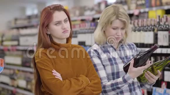 无聊的红发白种人女人等待女性朋友在商店里选择红白葡萄酒年轻迷人视频的预览图