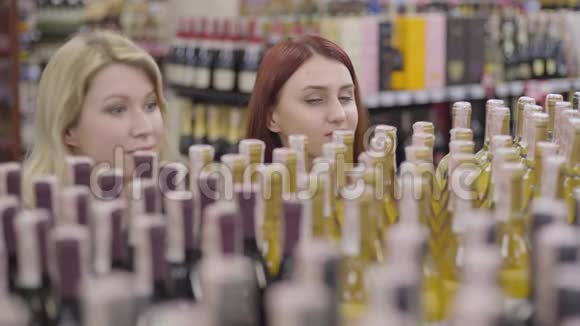 年轻的红发女人带着绿色的眼睛看着货架上的酒像一个开朗的金发朋友加入她的行列快乐的成人视频的预览图