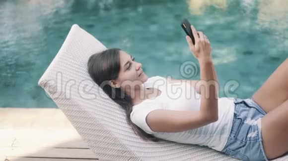 女人躺在游泳池附近的日光浴床上用电话工作输入信息视频的预览图