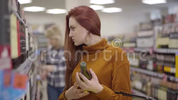 侧面看年轻美丽的红发女人检查货架与酒精和一瓶红酒在手中金发碧眼视频的预览图