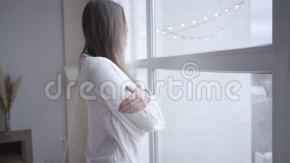 睡眼的年轻白种人女人早上打哈欠看着窗外从侧面看漂亮的深色头发视频的预览图