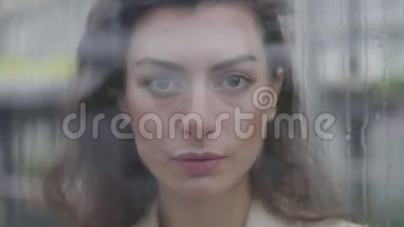 焦点从站在玻璃后面的迷人白种女人脸上的雨滴到触摸窗户视频的预览图