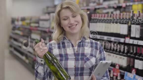一位金发碧眼的白人女性手持一瓶白葡萄酒和平板电脑对着镜头微笑年轻快乐视频的预览图