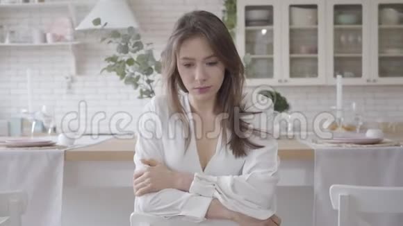美丽的白人女孩坐在时尚的厨房里思考的肖像忧郁的黑发白衣女青年视频的预览图