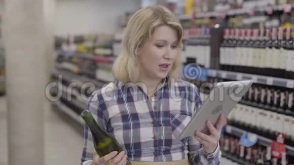 年轻开朗的白种人女人比较平板电脑上的信息和酒瓶上的标签积极的金发女郎选择视频的预览图