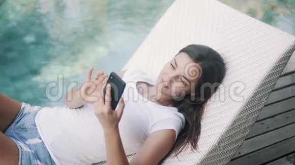 女自由职业者躺在游泳池附近的日光浴床上用电话工作输入信息视频的预览图