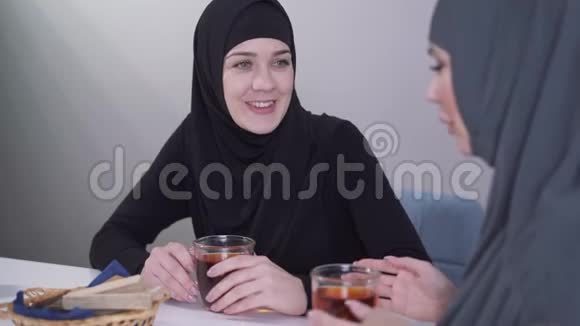 年轻穆斯林妇女戴着黑色头巾微笑着与朋友交谈的特写照片两个女人在室内交流视频的预览图