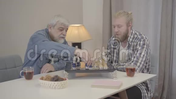 少年和年长男子在室内下棋的肖像白种人成年的儿子和成熟的父亲共度快乐的夜晚视频的预览图