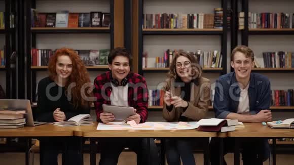 两个年轻的男学生和两个欧洲女学生坐在桌旁图书馆里放着书和笔记本电脑兴高采烈视频的预览图