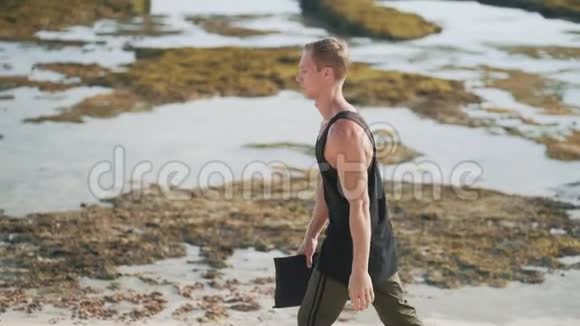 侧景手拿瑜伽垫的孤独男子沿着沙滩漫步视频的预览图