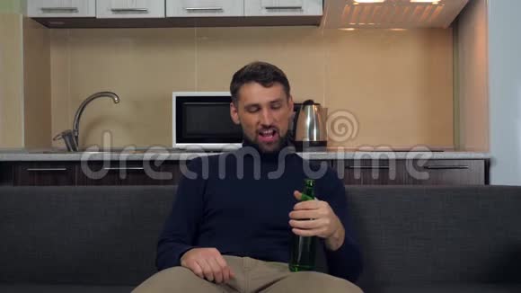 一个穿着深蓝色套衫和灰色裤子的年轻人坐在沙发上喝着啤酒微笑着看电视上的娱乐节目视频的预览图