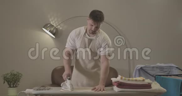 在家里熨衣服的白种人严肃的中镜头穿着围裙的帅哥履行家务视频的预览图