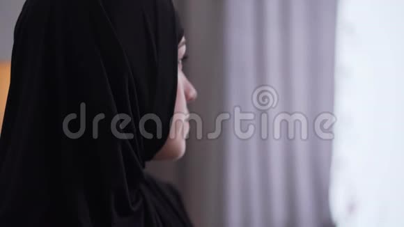 戴着头巾的漂亮心烦意乱的女人的特写脸看着窗外的家穆斯林妇女悲伤的肖像视频的预览图