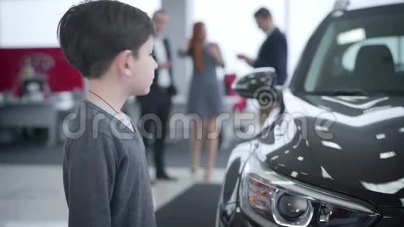 一个可爱的白人小男孩站在新车旁回头看着父母和商人交谈可爱的视频的预览图