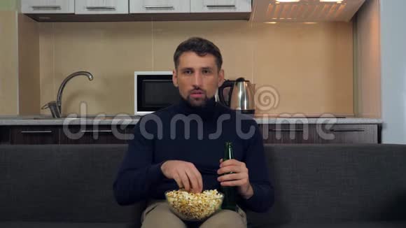 一个穿着深蓝色套衫和灰色裤子的年轻人坐在沙发上吃爆米花喝啤酒看电视上的娱乐节目视频的预览图