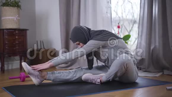 侧视苗条的穆斯林妇女弯着腰在瑜伽垫上试图用手达到脚尖性感的年轻女子视频的预览图