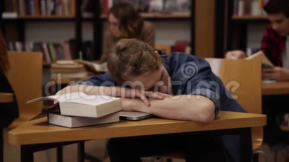 年轻的欧洲学生在大学图书馆或班级睡在桌子上上面有一堆书同学和书架视频的预览图
