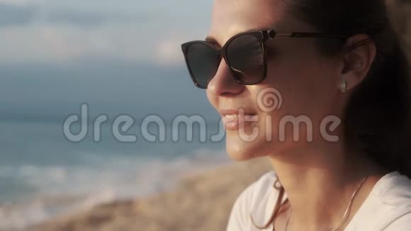 美丽微笑的女人戴着太阳镜在日落时看着大海的肖像视频的预览图