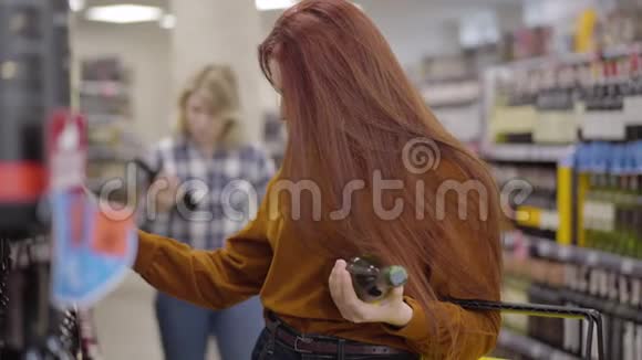 漂亮的红发女人选择超市货架上的饮料作为她的金发朋友带着一瓶葡萄酒来年轻女孩视频的预览图