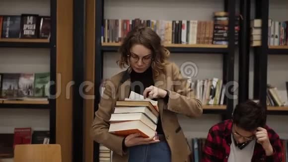 一个穿着棕色夹克和眼镜的女孩拿着一堆书从书架上拿下来的慢镜头视频的预览图