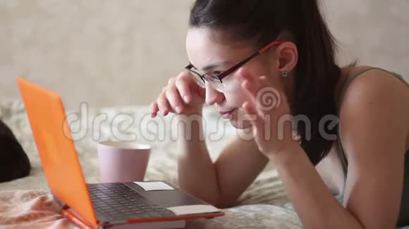 年轻漂亮的女孩正在用她的笔记本电脑工作躺在家里的床上一个年轻可爱的女孩躺在床上打字视频的预览图