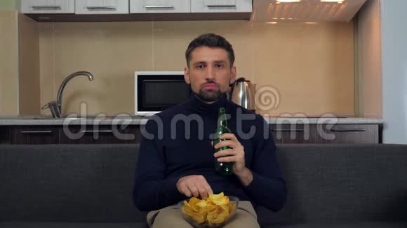 一个穿着深蓝色套衫和灰色裤子的年轻人坐在沙发上吃薯片喝啤酒看电视上的娱乐节目视频的预览图