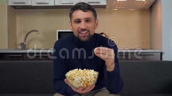 一个年轻人坐在沙发上吃透明玻璃盘子里的爆米花微笑着看电视上的娱乐节目视频的预览图