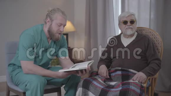 男性白种人志愿者穿着护士制服阅读书籍到盲人高级退休人员室内有同情心的男人晚上视频的预览图