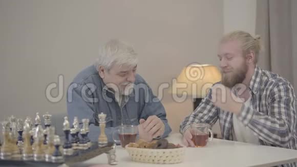 白种人成年男子坐在桌子旁在室内聊天的肖像有胡子的儿子周末和年长的儿子在一起视频的预览图