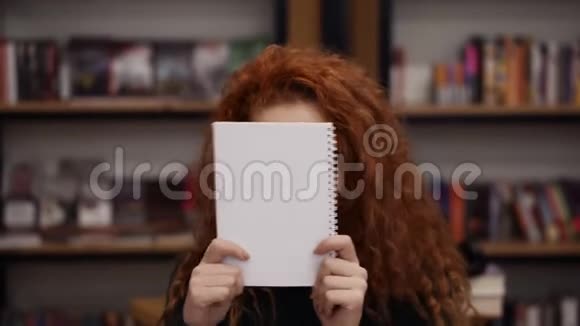 幸福的红头女孩用记事本藏着脸的肖像然后微笑地站在书架背景上青年文化视频的预览图