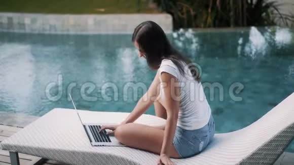 欢乐女孩的侧景在笔记本电脑上工作坐在游泳池附近的沙发上视频的预览图